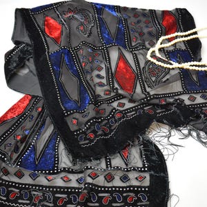May include: A rectangular scarf featuring a sheer gray base with black velvet borders and fringe. The fabric is decorated with red and blue diamond-shaped patches, paisley motifs, and small silver-toned beads. A string of white faux pearls rests on top.