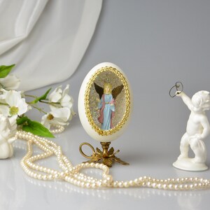Puede incluir: Un huevo de porcelana blanca con un borde dorado y un interior lleno de purpurina, que presenta una figurita de &aacute;ngel en miniatura en su interior. El huevo se muestra en un soporte de metal dorado con tres patas. Tambi&eacute;n se muestra una figurita de querub&iacute;n de porcelana blanca, sosteniendo un anillo en su mano. Un collar de perlas blancas est&aacute; en primer plano.