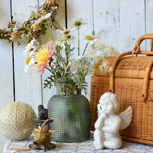 May include: A green textured glass vase filled with white daisies and a pink dahlia, a white ceramic angel figurine, a round fabric-covered ball, a small brass bell on a wooden spool, and a woven wicker basket on a white lace-covered table.