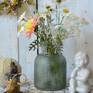 May include: A clear green glass vase with a textured hobnail pattern holds a bouquet of white daisies, a pink and yellow dahlia, and silver-toned dusty miller foliage. The vase sits alongside a small white ceramic angel figurine and a metal bell.