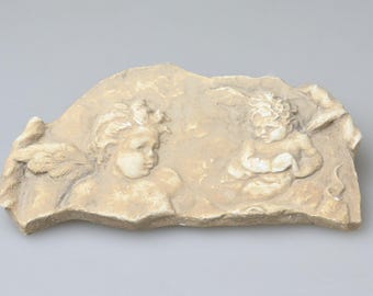 Puede incluir: Una placa de relieve de yeso beige de forma semicircular que presenta dos querubines esculpidos con alas. Las figuras están representadas con un acabado suave y texturizado con detalles en relieve que muestran sus rostros, cabello y alas.