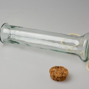 May include: A clear glass vase with a wide opening and a cork stopper. A strand of pearls is draped over the vase. The vase is lying on its side on a white surface. The vase is approximately 10 inches long.