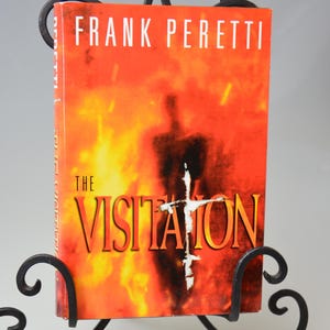 May include: A book titled "The Visitation" by Frank Peretti, displayed in a black wrought iron stand. The book cover features a fiery orange and red background with a silhouette and a white cross.