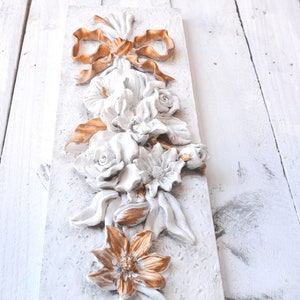 May include: A white and gold floral wall decor with a 3D design. The flowers are made of plaster and painted in a white and gold color scheme. The flowers are arranged in a cascading design, with a bow at the top.