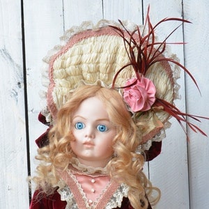 May include: A porcelain doll wearing a red velvet dress with a lace trim, a pink satin skirt, and a white hat with a pink flower. The doll has blonde hair and blue eyes.