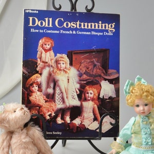 May include: A book titled "Doll Costuming: How to Costume French & German Bisque Dolls" by Jean Seeley is displayed on a black metal stand. A teddy bear and a porcelain doll are sitting in front of the book.