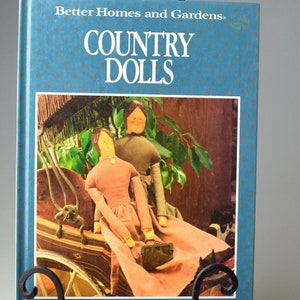 Puede incluir: Un soporte para libros de metal negro que sostiene un libro titulado "Better Homes and Gardens Country Dolls". La portada del libro muestra una foto de dos muñecas sentadas en un carruaje. Una pequeña caja de madera con un diseño de corazón está en el fondo.