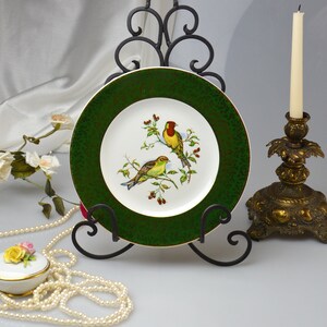 May include: A green and white porcelain plate with a gold rim and a painted design of two birds on a branch. The plate is displayed on a black metal stand.