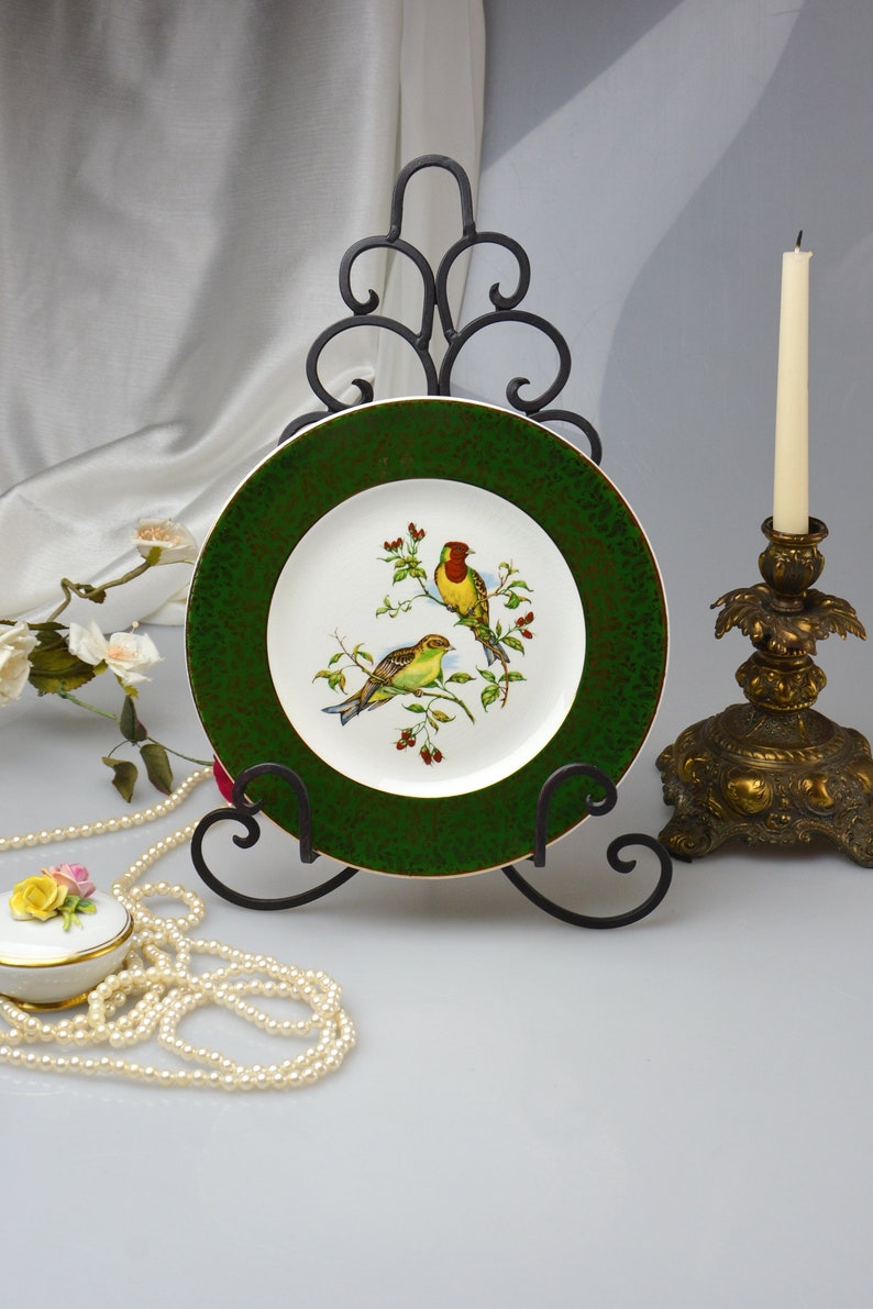 May include: A green and white porcelain plate with a gold rim and a painted design of two birds on a branch. The plate is displayed on a black metal stand.