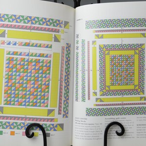 May include: Two open pages of a book showing quilt assembly diagrams. The diagrams use colourful geometric patterns, including triangles and squares in yellow, pink, green, blue, and orange. The pages are held open by a black metal book stand.