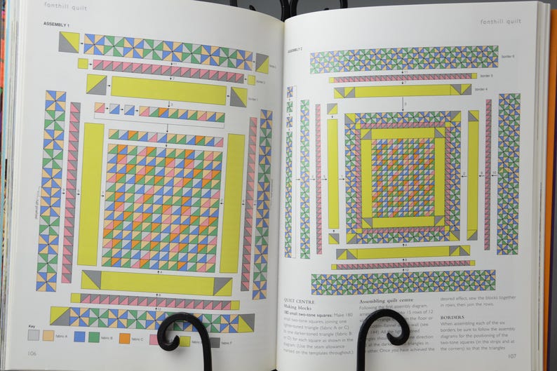 May include: Two open pages of a book showing quilt assembly diagrams. The diagrams use colourful geometric patterns, including triangles and squares in yellow, pink, green, blue, and orange. The pages are held open by a black metal book stand.