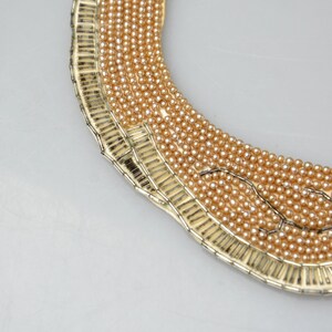 May include: A gold-toned choker necklace with a wide, rounded shape. The necklace is made of small, round beads that are arranged in a pattern. The beads are a light gold color.
