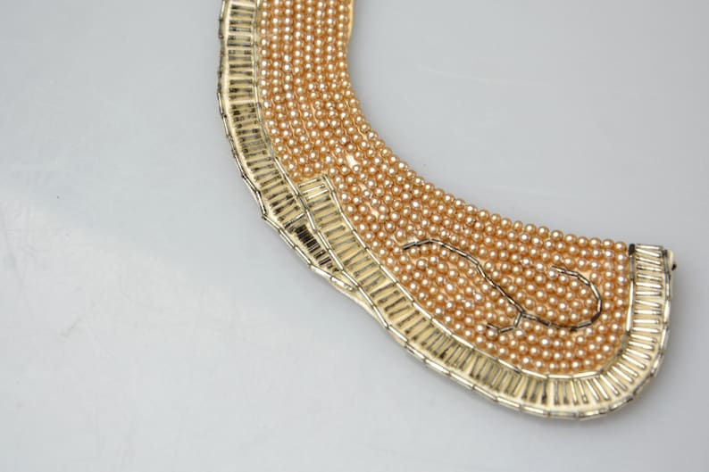 May include: A gold-toned choker necklace with a wide, rounded shape. The necklace is made of small, round beads that are arranged in a pattern. The beads are a light gold color.