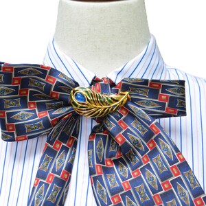 May include: A white dress shirt with thin blue and pink vertical stripes, featuring a large, tied bow necktie made of dark blue fabric with a repeating geometric pattern in red and gold, secured by a gold-toned peacock feather brooch with a blue stone.