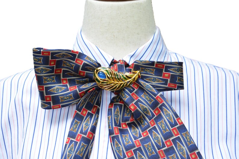 May include: A white dress shirt with thin blue and pink vertical stripes, featuring a large, tied bow necktie made of dark blue fabric with a repeating geometric pattern in red and gold, secured by a gold-toned peacock feather brooch with a blue stone.