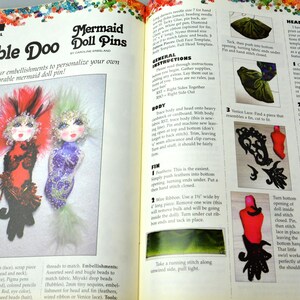May include: A craft book page with instructions on how to make mermaid doll pins. The page features a colorful illustration of three mermaid doll pins with different embellishments. The text includes the title "Bubble Doo Mermaid Doll Pins" and instructions for making the doll pins.