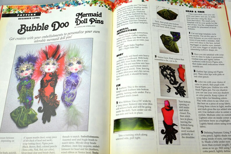 May include: A craft book page with instructions on how to make mermaid doll pins. The page features a colorful illustration of three mermaid doll pins with different embellishments. The text includes the title "Bubble Doo Mermaid Doll Pins" and instructions for making the doll pins.