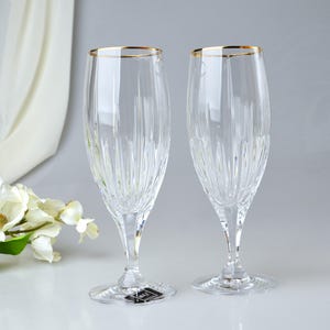 Pu&ograve; includere: Due fl&ucirc;te da champagne trasparenti con bordo dorato. I bicchieri hanno un design verticale inciso e poggiano su una base trasparente. Lo sfondo &egrave; bianco e grigio chiaro con un rametto di fiori bianchi.