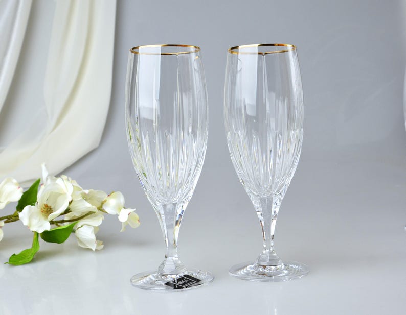 Pu&ograve; includere: Due fl&ucirc;te da champagne trasparenti con bordo dorato. I bicchieri hanno un design verticale inciso e poggiano su una base trasparente. Lo sfondo &egrave; bianco e grigio chiaro con un rametto di fiori bianchi.