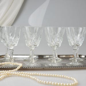May include: A set of six clear crystal wine glasses with a detailed, etched design, displayed on a silver-toned serving tray. A strand of faux pearls is draped in front of the tray. The glasses are approximately 7 inches tall.