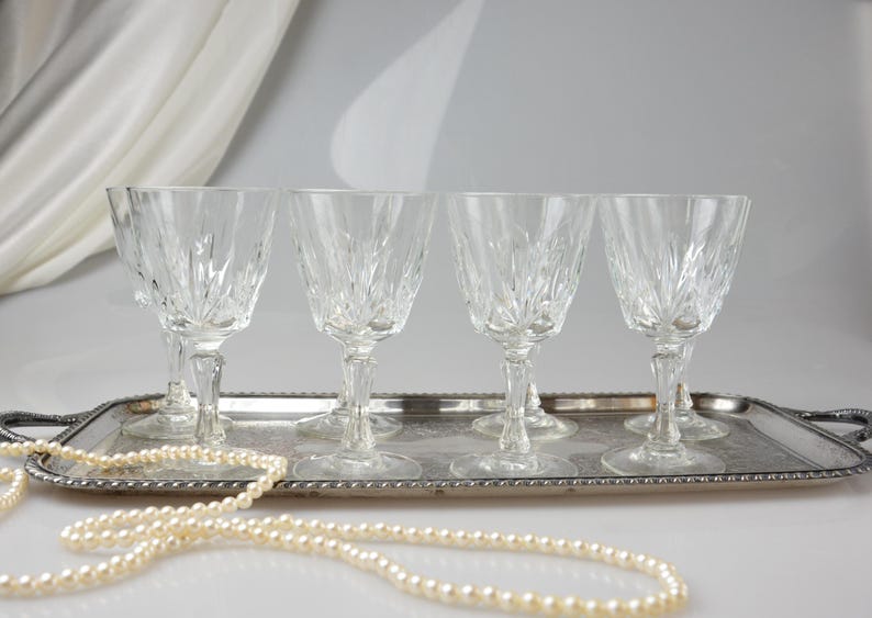 May include: A set of six clear crystal wine glasses with a detailed, etched design, displayed on a silver-toned serving tray. A strand of faux pearls is draped in front of the tray. The glasses are approximately 7 inches tall.