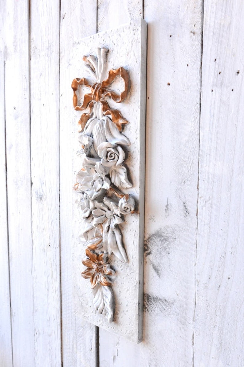 May include: A white rectangular wall plaque with a 3D floral design. The flowers are white with gold accents and a gold bow at the top.