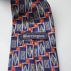 May include: A necktie featuring a repeating geometric pattern of blue rectangles, gold diamond shapes with blue floral motifs, and small red squares. A rectangular fabric label with the text Barrington is stitched across the center.