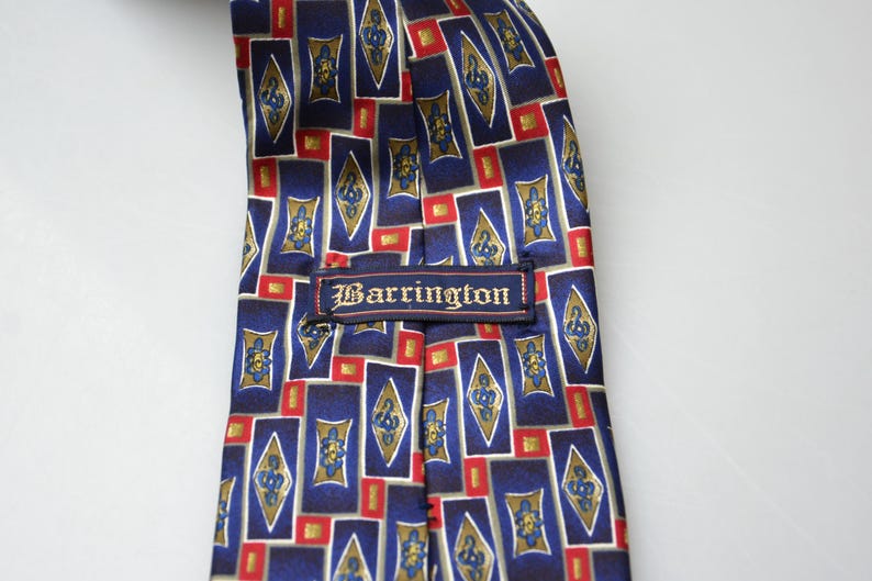 May include: A necktie featuring a repeating geometric pattern of blue rectangles, gold diamond shapes with blue floral motifs, and small red squares. A rectangular fabric label with the text Barrington is stitched across the center.