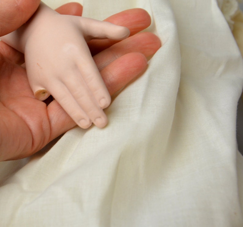 May include: A close-up of a porcelain doll's hand resting on a white fabric surface. The doll's hand is pale and has delicate fingers.