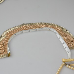 May include: A gold and cream beaded collar necklace with a measuring tape wrapped around it. The necklace is shaped like a crescent moon.
