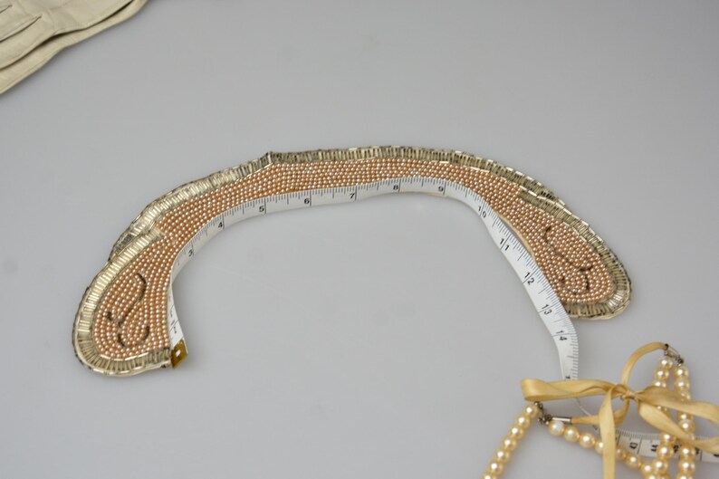 May include: A gold and cream beaded collar necklace with a measuring tape wrapped around it. The necklace is shaped like a crescent moon.