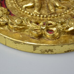 May include: A gold-colored round plaque with a raised design of figures and a wreath. The plaque has a red background and is decorated with gold leaf.