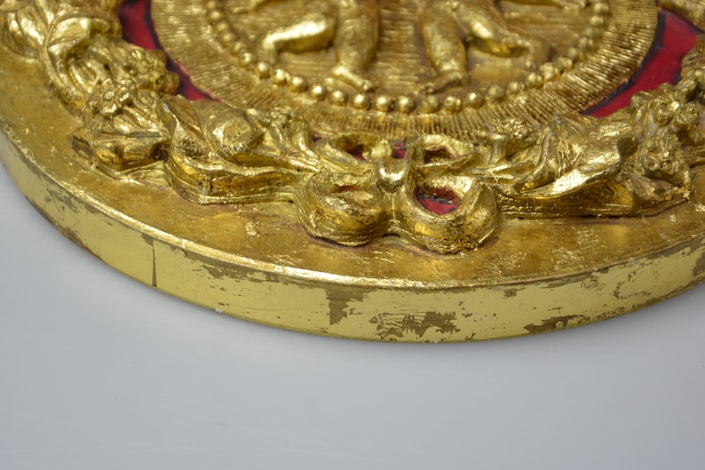May include: A gold-colored round plaque with a raised design of figures and a wreath. The plaque has a red background and is decorated with gold leaf.