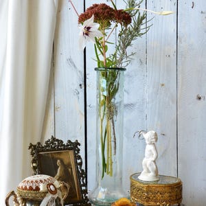 K&ouml;nnte beinhalten: Ein Stillleben mit einer hohen Glasvase mit Blumen, einem gerahmten Vintage-Foto, einer Schmuckschatulle und einer kleinen wei&szlig;en Cherubfigur. Die Gegenst&auml;nde sind auf einem mit Spitze bedeckten Tisch vor einem wei&szlig;en Holzhintergrund angeordnet.