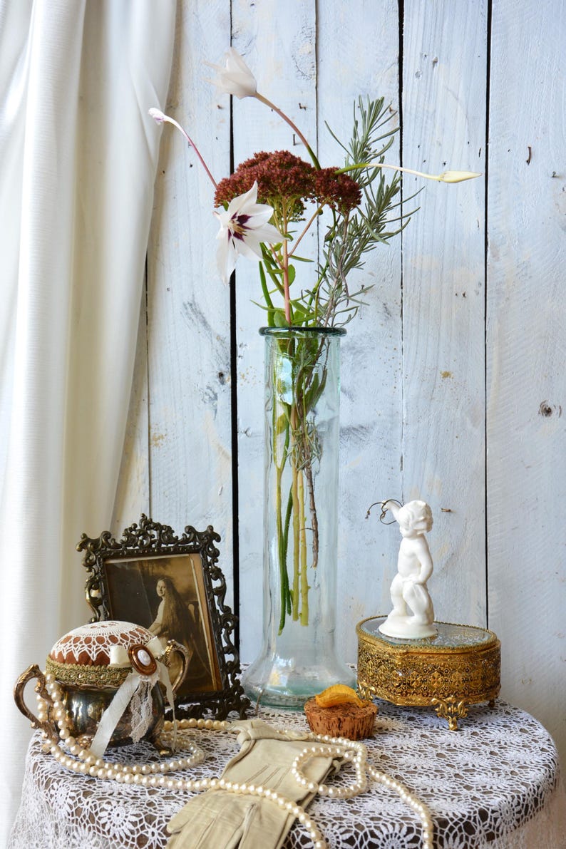 K&ouml;nnte beinhalten: Ein Stillleben mit einer hohen Glasvase mit Blumen, einem gerahmten Vintage-Foto, einer Schmuckschatulle und einer kleinen wei&szlig;en Cherubfigur. Die Gegenst&auml;nde sind auf einem mit Spitze bedeckten Tisch vor einem wei&szlig;en Holzhintergrund angeordnet.