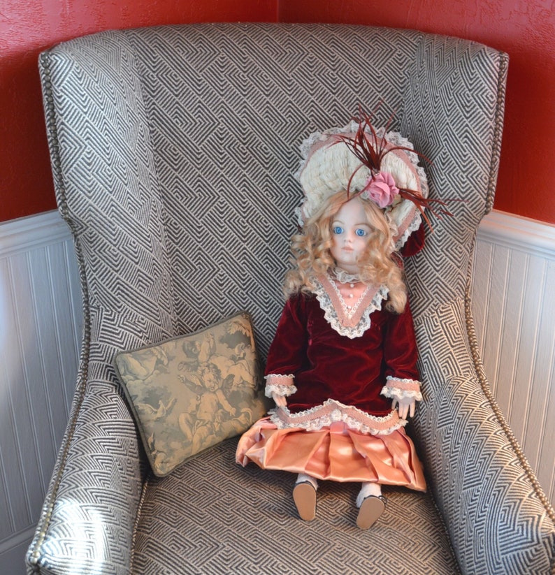 May include: A porcelain doll wearing a pink and white dress with a red velvet jacket sits in a gray armchair with a patterned fabric. The doll has blonde hair and blue eyes and is wearing a white hat with a pink flower.
