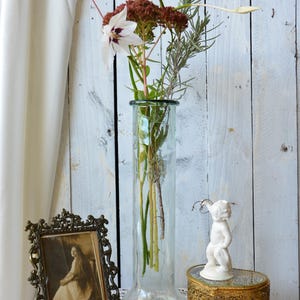 K&ouml;nnte beinhalten: Eine klare Glasvase enth&auml;lt ein Blumenarrangement mit wei&szlig;en und burgunderfarbenen Bl&uuml;ten. Ein Vintage-Fotorahmen, eine kleine goldene Schmuckschatulle und eine wei&szlig;e Cherubfigur stehen auf einem mit Spitze bedeckten Tisch. Ein Korken und ein Bl&uuml;tenblatt sind ebenfalls vorhanden.