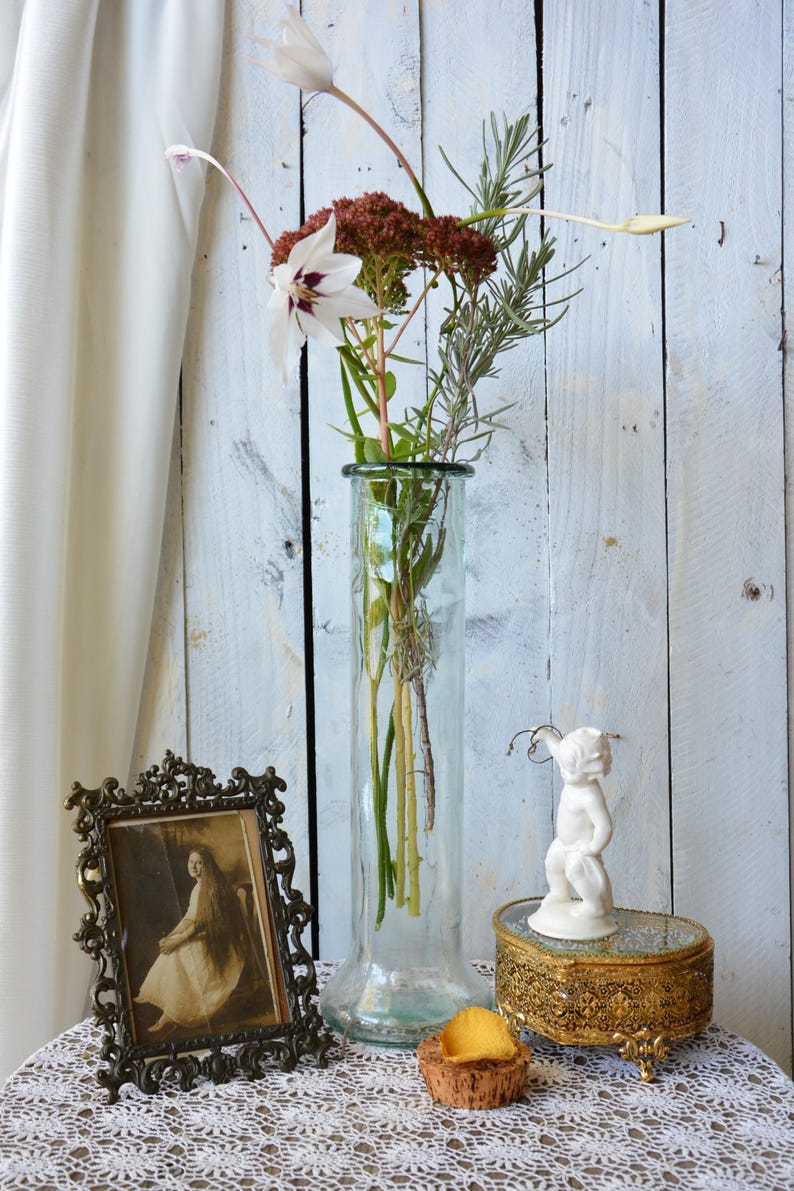 K&ouml;nnte beinhalten: Eine klare Glasvase enth&auml;lt ein Blumenarrangement mit wei&szlig;en und burgunderfarbenen Bl&uuml;ten. Ein Vintage-Fotorahmen, eine kleine goldene Schmuckschatulle und eine wei&szlig;e Cherubfigur stehen auf einem mit Spitze bedeckten Tisch. Ein Korken und ein Bl&uuml;tenblatt sind ebenfalls vorhanden.