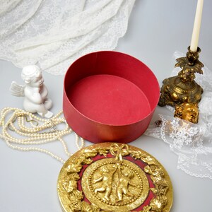May include: A round, red box with a gold lid featuring two cherubs holding a ribbon. The lid is decorated with gold leaf and intricate floral designs.