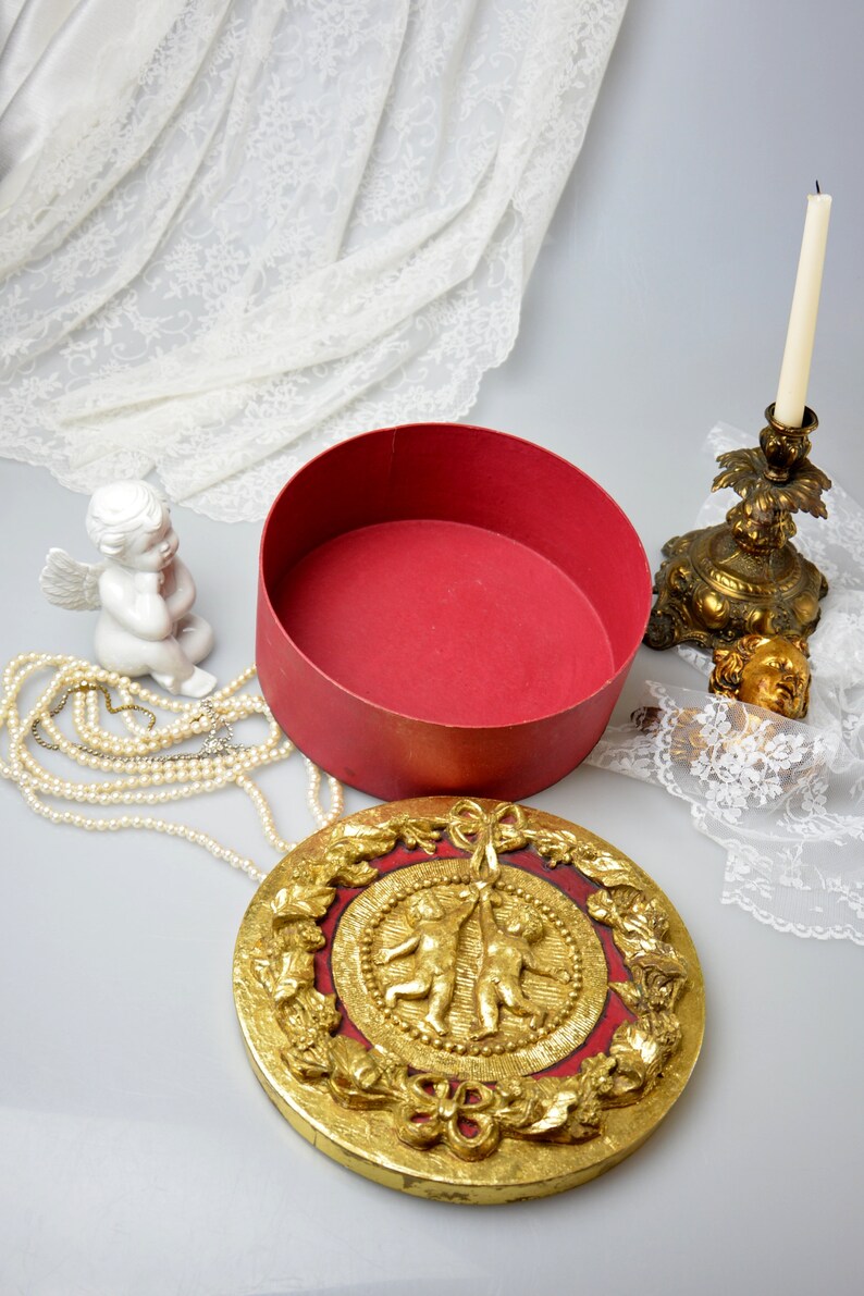 May include: A round, red box with a gold lid featuring two cherubs holding a ribbon. The lid is decorated with gold leaf and intricate floral designs.