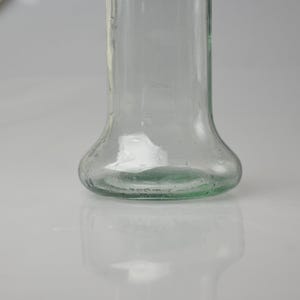 K&ouml;nnte beinhalten: Eine klare Glasflasche mit bauchigem Boden und einem Korken. Die Flasche ist leer und reflektiert das Licht, mit einem leichten Gr&uuml;nstich am Boden. Der Korken hat eine hellbraune Farbe.