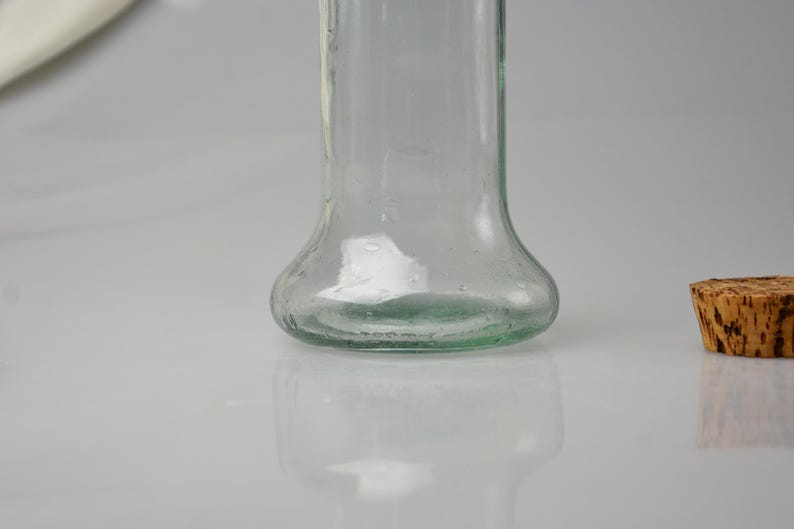 K&ouml;nnte beinhalten: Eine klare Glasflasche mit bauchigem Boden und einem Korken. Die Flasche ist leer und reflektiert das Licht, mit einem leichten Gr&uuml;nstich am Boden. Der Korken hat eine hellbraune Farbe.
