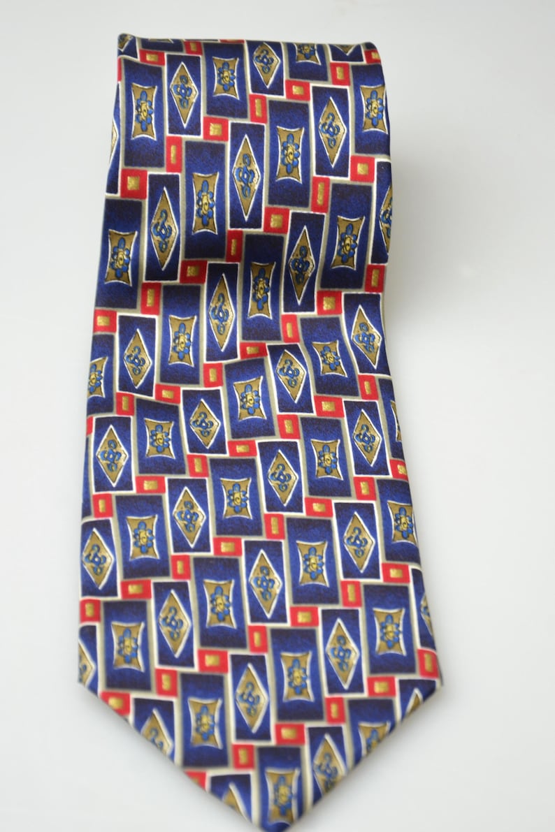 May include: A necktie featuring a repeating geometric pattern of dark blue rectangles and gold diamond shapes. Each diamond contains a small blue floral motif, and the design is accented with small red squares interspersed between the shapes.