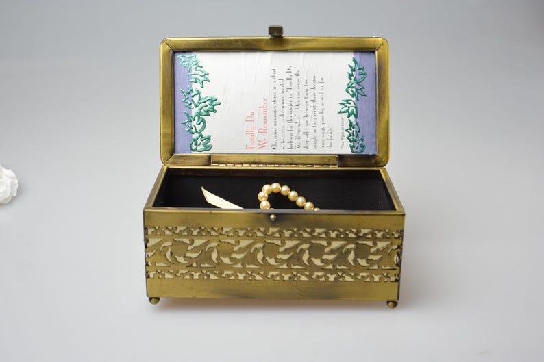 Op de afbeelding: Een rechthoekige messing sieradendoos met een open scharnierend deksel. De doos heeft een decoratief bloemmotief en kleine voetjes. Binnenin zijn een parelketting en een kleine kaart zichtbaar. Het deksel toont tekst en bloemaccenten.