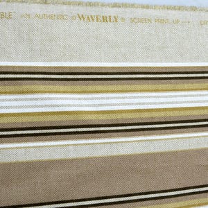 Puede incluir: Tela beige con rayas horizontales en tonos marr&oacute;n, negro, blanco y dorado. El texto "STRIPE ENSEMBLE" y otro texto est&aacute;n impresos en la parte superior. La tela es probablemente para tapicer&iacute;a o decoraci&oacute;n del hogar.