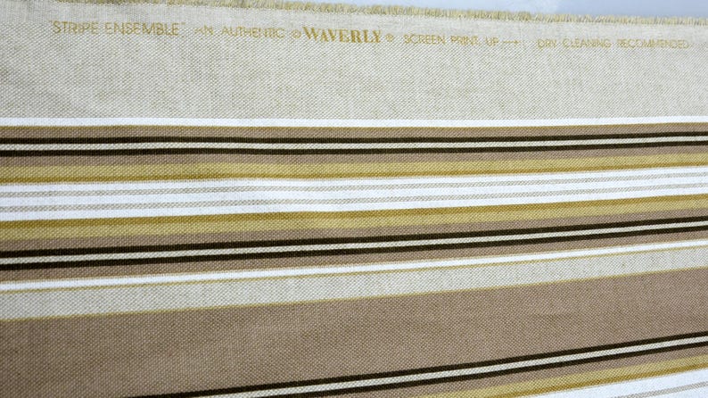 Puede incluir: Tela beige con rayas horizontales en tonos marr&oacute;n, negro, blanco y dorado. El texto "STRIPE ENSEMBLE" y otro texto est&aacute;n impresos en la parte superior. La tela es probablemente para tapicer&iacute;a o decoraci&oacute;n del hogar.