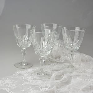 May include: Four clear crystal wine glasses with intricate etched designs. The glasses are arranged on a white lace tablecloth, creating an elegant and refined aesthetic. The stems and bases are also detailed.