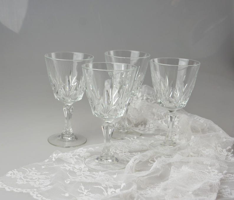 May include: Four clear crystal wine glasses with intricate etched designs. The glasses are arranged on a white lace tablecloth, creating an elegant and refined aesthetic. The stems and bases are also detailed.
