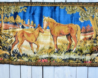 Può includere: Un arazzo raffigurante una scena campestre con due cavalli. I cavalli sono di varie tonalità di marrone e beige, su uno sfondo di cielo blu e un paesaggio con alberi e un piccolo edificio. L'arazzo ha un bordo decorativo.