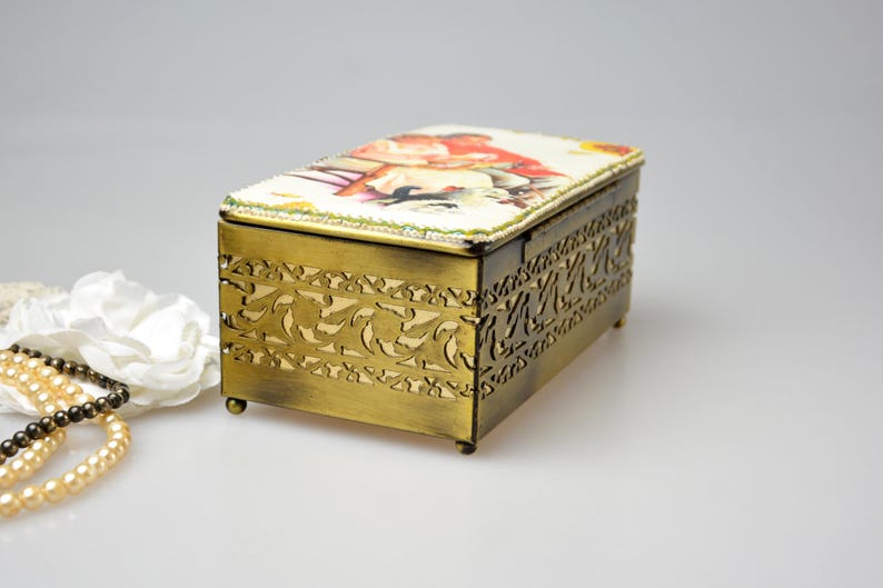 May include: Rectangular antique brass jewelry box with a decorative floral design on the sides. The lid features a colorful illustration. The box is set on small feet and has a beaded trim.